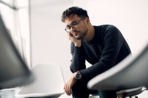 Young worried man sitting in a chair and thinking while waiting a group therapy to start.