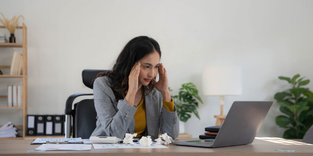 Tired business woman in stress works at a laptop while sitting at a table at home and holds her hand on her temples, migraine attack. Freelance, work from home.